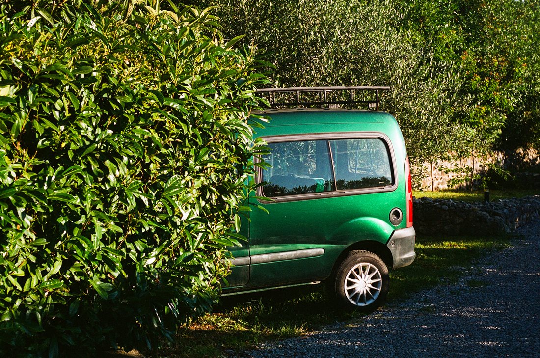 Ardèche, arrière de voiture verte type Berlingot à demi cachée par végétation, allée de gravier, arbres, pelouse, mur